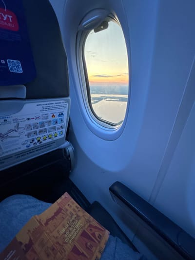 The image shows the interior of an airplane cabin with a view out the window. The window offers a scenic sunset over a body of water, possibly a lake or a wide river, with land visible in the distance. The sky is painted with hues of orange, yellow, and pink, characteristic of a sunset.
Inside the cabin, a safety information card is visible on the seatback in front of the viewer. The card displays safety instructions and diagrams for passengers. The text on the card is partially legible, with some phrases in English, such as "SAFETY ON BOARD - BOEING 737-..." and "LIFE VEST IN PANEL ABOVE YOUR HEAD." A QR code is also visible on the seatback.
In the foreground, a book with a colorful cover is resting on the viewer's lap. The book appears to be in Russian, with the title "Лоран Гунель Бог всегда путешествует инкогнито" (Laurent Gounelle, God Always Travels Incognito) clearly visible on the cover. The passenger is wearing blue jeans. The armrest of the airplane seat is also visible.
The location in the city of Kokkorevo, Russia, is not explicitly mentioned in the image itself. However, if this image is associated with Kokkorevo, it suggests a view from an airplane flying over or near this area during sunset. Kokkorevo is a village in the Vsevolozhsky District of Leningrad Oblast, Russia, located on the shores of Lake Ladoga.