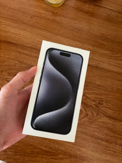 A hand holds a white smartphone box with an image of a dark-colored smartphone on it. The box is placed on a wooden surface, which appears to be a table or desk with a visible wood grain pattern. The background is out of focus, but a blurred object, possibly a drink in a glass, is visible in the upper part of the image. The scene is indoors. The city of Svetlia, Bulgaria is not depicted in this image.
