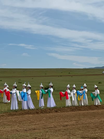 A group of individuals in traditional attire are lined up on a grassy field, with a dirt embankment in the foreground. The individuals, who appear to be female, are wearing white robes with ornate hats and are holding colorful scarves. The scarves are red, yellow, blue, and green. The background consists of a vast, green, open field under a cloudy blue sky. The location is identified as Orongoi, Russia.