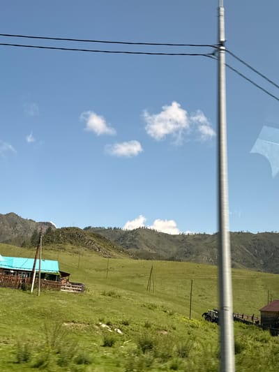 This is a photograph taken from a moving vehicle, likely a car, looking out at a rural landscape in the vicinity of Tuekta, Russia. The scene is dominated by a vast, green, rolling pasture under a bright blue sky with scattered white clouds. In the mid-ground, a small wooden house with a distinctive turquoise roof and a wooden fence is visible on the left side of the frame. A wooden pole, likely a utility pole, stands in front of the house. Further into the field, to the right of the house, a blue tractor is parked on a slight incline. In the background, rugged, tree-lined mountains recede into the distance. Power lines are strung across the top of the image, and a tall, metallic utility pole runs vertically along the right side of the frame, obscuring part of the view. The overall impression is one of a serene, natural environment in a remote, possibly agricultural, setting.