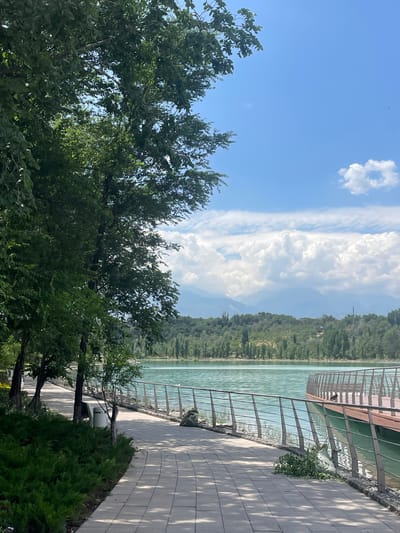 The image depicts a scenic view of a lakeside promenade on a sunny day in Almaty, Kazakhstan. The promenade, paved with light-colored square tiles, curves along the edge of a tranquil turquoise lake. A silver metal railing runs along the lakeside, separating the path from the water. Lush green trees with dense foliage line the left side of the path, casting dappled shadows on the ground. In the foreground, neatly trimmed green bushes are visible along the edge of the path.
Further along the promenade, a wooden deck juts out over the water, also equipped with a metal railing. A small bird is perched on this railing. The lake's surface is calm, reflecting the bright blue sky dotted with fluffy white clouds. In the background, a range of tree-covered hills and distant, hazy mountains form a picturesque backdrop. The overall atmosphere is serene and inviting, suggesting a popular recreational spot in Almaty.