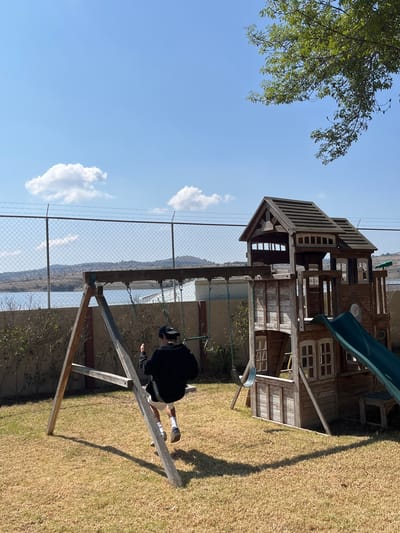 A photo shows a person on a swing set in what appears to be the backyard of a home in El Oasis Valsequillo, Mexico. The individual is wearing a black top, light-colored shorts, socks, and shoes. They seem to be in motion, suggesting the swing is in use. To the left is a wooden playhouse with a slide attached. In the background, a wire fence separates the yard from a distant landscape, likely a body of water with hills beyond. The sky is blue with scattered clouds. The yard is covered in dry grass. A tree is partially visible on the left side of the image.
