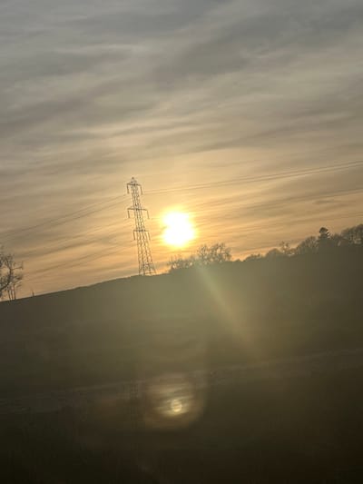 The image depicts a rural landscape near Fowlis Easter, United Kingdom, at what appears to be sunrise or sunset. The scene is heavily backlit by a low-positioned sun, slightly right of the upper center, which creates a strong glow and visible lens flare effects in the lower portion of the frame, including a circular, secondary light source in the lower center. The sky is overcast with thin, horizontally stratified clouds, appearing warm yellow and orange near the horizon, transitioning to lighter grey further up.
A large, steel lattice power line pylon stands prominently on a gentle, dark-colored hillside, slightly to the left of the image's center, with multiple power lines extending horizontally from it across the sky. The terrain slopes upwards from the left and foreground towards the right and background. The foreground and midground consist of dark, indistinct ground cover, possibly fields or natural vegetation, with a subtle, lighter-toned horizontal track or path visible across the lower midsection. A line of bare deciduous trees is partially visible on the far left, while a denser treeline, comprising both deciduous and evergreen trees, crests the hill in the background, silhouetted against the bright sky. No individuals, animals, or vehicles are discernible within the frame.