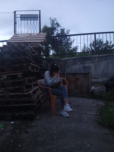 This image depicts an outdoor scene in Zemen, Bulgaria, at what appears to be dusk or early morning under an overcast sky. The central subject is a young woman seated on a bright orange plastic chair. She wears a light-colored long-sleeved top, ripped blue jeans, and white athletic shoes, with her hair tied back in a ponytail. Her face is partially obscured by her right hand, while her left hand holds a dark smartphone, suggesting she is either engrossed in her phone or experiencing a moment of contemplation or distress.
To the woman's left, a large, dark pile of wooden pallets is stacked high. In the upper left corner, another structure made of wooden pallets rises, leading to a decorative black metal frame with diagonal cross-patterned bars and a rectangular, unidentifiable grey object suspended within it. A dark metal railing runs across the upper background, separating the foreground from distant trees and foliage. Behind the woman, a concrete wall extends, with a rusty metal sheet leaning against it on the right. The ground is an uneven mix of dirt and gravel, with some scattered debris, including a small green bottle and pieces of paper. The overall setting appears to be an unkempt, industrial, or neglected outdoor area. No discernible text is visible in the image.