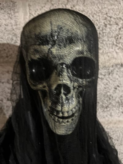 The image displays a close-up, front-facing perspective of a decorative skull head, positioned against a light-colored, textured wall in Vieux, France. The skull itself is off-white or beige, featuring dark grey or black painted details that simulate cracks and shadows, particularly around the eye sockets, nasal cavity, and the grinning teeth. A fine, black mesh or net fabric overlays the entire skull, creating a visible grid pattern across its surface. This black fabric also drapes downwards from the skull, forming a shroud-like covering that extends out of the bottom of the frame. The eye sockets contain dark, reflective black circular inserts. The background wall exhibits a rough, irregular texture consistent with stone or stucco. No individuals are present, and no actions or interactions are depicted.
