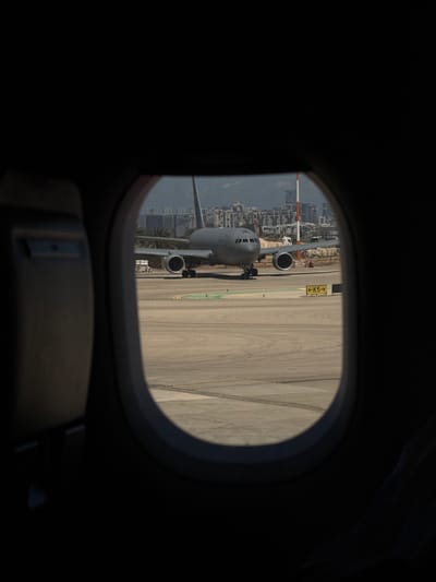 The image shows a view through an airplane window. Another, larger, gray airplane with two visible engines is seen on the tarmac, near a city skyline, which places the location in Sdot Dan Regional Council, Israel. The wing of the plane can be seen in the background. A taxiway direction sign is visible near the plane. The interior of the airplane from which the image is taken is dark.