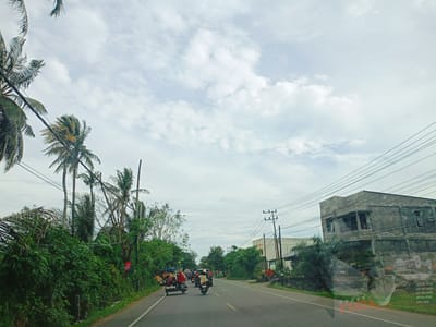 A view from a vehicle traveling along a two-lane road in Lampisang, Indonesia. The sky is overcast with white clouds. The road is gray asphalt. The left side of the road is bordered by dense, green vegetation, including palm trees. On the right side of the road, there are buildings, including a gray, two-story structure under construction. Power lines crisscross above the road. Several motorbikes and a sidecar motorcycle are visible on the road ahead, traveling in the same direction as the camera.
