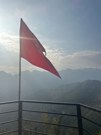 The scene, located in Minh Tân Commune, Vietnam, displays a red flag with a yellow five-pointed star, consistent with the Vietnamese national flag, flying from a bamboo pole. The flag exhibits a tear on its trailing right edge. Bright sunlight from the upper left casts a lens flare across the flag. The background consists of multiple layers of vegetated mountains receding into a distant atmospheric haze. Beneath the mountains, a valley shows faint indications of human settlement and winding paths. A dark metal railing with horizontal bars occupies the lower foreground, suggesting an elevated vantage point. No human figures are present in the image.
