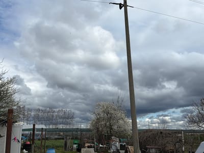 The image displays an outdoor, semi-rural scene situated in Liahovo, Bulgaria, under an overcast sky. The sky is predominantly covered by extensive grey and white cumulus clouds, with isolated sections of lighter blue visible in the upper left and along the right horizon.
A grey concrete utility pole, equipped with dark insulators and horizontal wires, occupies the right side of the foreground, extending vertically. To the left of this pole, a deciduous tree in full white blossom is prominent. Below the blooming tree, a cluttered area includes what appears to be discarded household appliances, specifically a washing machine and an air conditioner unit, along with wooden planks and assorted debris. A green crate labeled "TUBORG" is visible on the ground towards the center-left. On the mid-right, a large accumulation of bare, tangled branches rests behind a wooden structure.
The left foreground features a structure with a white door, near which pink and black clothes pegs are attached to a line. Rusty metal poles and a blue barrel are also present in this area. Beyond these elements, the middle ground is characterized by green vegetation and multiple white and green structures resembling greenhouses or agricultural tunnels. Behind these structures, a horizontal line of tall, bare deciduous trees with slender trunks extends across the frame. The distant background reveals additional bare trees and a faint utility tower against a lighter patch of sky on the right horizon. No individuals are present within the scene. The combination of blooming trees and bare branches indicates an early spring season.