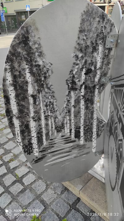 The image captures an outdoor scene featuring a round, oval-shaped artwork painted with a black and white depiction of a birch tree-lined path. The painting is mounted on a vertical metallic structure, with visible bolts and brackets attaching it. To the right of the painting, a portion of another metallic object with a circular design, possibly a clock face, is visible. The artwork is situated on a cobbled street, with some sparse greenery growing between the stones. In the background, a building with windows and awnings, a traffic light, and a blue and white triangular sign are discernible, indicating an urban environment. The scene is illuminated by natural light. A timestamp "2026/7/8 18:32" is present in the lower right corner of the image. The style of the artwork and the surrounding architecture suggests it is located in a European city. Based on the date and general appearance, this artwork is likely a public installation in Iisalmi, Finland, possibly part of a seasonal art exhibition or permanent street art.