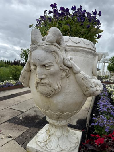 An ornately decorated, weathered stone urn, featuring a sculpted male face with a beard and horns, stands prominently in an outdoor setting. The urn is filled with vibrant purple flowers and lush green foliage. It is placed on a paved walkway with a patterned stone surface. Surrounding the urn are beds of purple and white flowers, as well as red foliage. In the background, a white pergola-like structure and trees are visible, suggesting a garden or park environment. The sky is overcast with clouds. The location is identified as Bruzdeilynas, Lithuania.