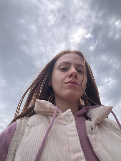 This is a close-up, low-angle, outdoor, selfie-style photograph of a young Caucasian woman. She has long, straight, light brown hair that is windswept and partially obscures her face. Her head is tilted slightly downwards, and she is looking directly at the camera with a neutral expression. She is wearing a purple hooded sweatshirt with a cream-colored corduroy vest over it. The vest has a ribbed texture and is fastened with white buttons. A purple drawstring is visible on the vest. The background consists of a cloudy, overcast sky. The location is identified as Son en Breugel, Netherlands.