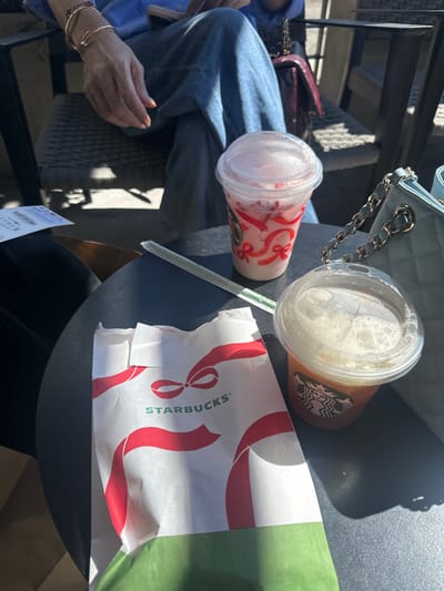 Two Starbucks cups with lids and a straw rest on a table next to a folded Starbucks paper bag. One cup is clear with red ribbon designs, while the other is partially visible with the Starbucks logo and a light-colored drink. A light blue quilted purse with a chain strap sits nearby. A person wearing a blue shirt and jeans is visible, with their hand adorned with gold bracelets. A small portion of a receipt can be seen on the table.