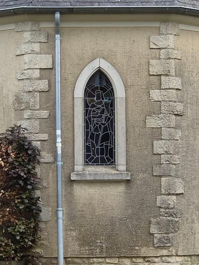 The image shows a section of a building's exterior, likely in Schengen, Luxembourg. A stained-glass window with a pointed arch is centered in the frame. Above and below the window are rows of rough-hewn, rectangular stone blocks that appear to be decorative elements integrated into the wall. A metal pipe runs horizontally across the wall, just below the window. Some shrubbery is present at the bottom of the frame. The building's facade is textured and appears to be constructed from stone or concrete. The overall color palette is muted, consisting mainly of grey, beige, and hints of brown.