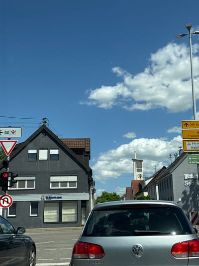 The image captures a sunny day in Nürtingen, Germany, as seen from a vehicle stopped at an intersection. The foreground is dominated by the rear of a silver Volkswagen Golf, partially obscuring the view of the road and crossing traffic. To the left, a black car is visible.

In the middle ground, a dark gray building with a pitched roof stands prominently. Its facade features three evenly spaced windows on the upper level and two larger windows on the lower level, all covered by white blinds. The lower level also has signage for "Dr. Koch Dr. Goth," indicating a dental practice. To the right of this building, a lighter-colored building with multiple windows is visible, with a sign reading "Münting Senner..."

The background reveals a clear blue sky with scattered white clouds. A modern street lamp with multiple lights stands to the right. A church tower with a clock face is visible above the buildings, and several directional signs are posted, including one pointing to "297 Kirchheim" and another to "FH-N" and "Waldhafen." Greenery is present, suggesting a town or city environment. The traffic light at the intersection is red.