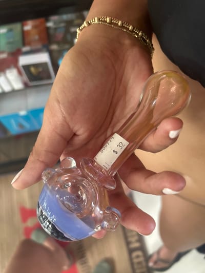 A person is holding a glass pipe in their hand. The pipe is clear with a blue and white design and has a price tag attached to it that reads "$32." The person is wearing a gold bracelet. The image was taken in Cobano District, Costa Rica, at a latitude of 9.635 and a longitude of -85.158. The environment is not clearly visible in the image, but it appears to be a store or shop. The subject is the glass pipe, and the action is holding. The person is interacting with the pipe, and the environment is the store or shop.