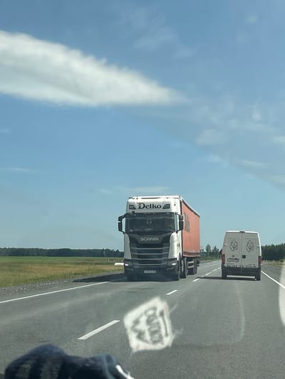 This image depicts a view from a vehicle traveling on a highway, likely in the vicinity of Iliushkinskaya, Russia, given the context provided. The sky is a clear blue with a few wispy white clouds. On the highway, a large white semi-truck with an orange trailer, branded "Delko" and "SCANIA," is in the left lane. In the right lane, a white van with two cartoon bear logos on its rear doors is traveling ahead of the semi-truck. The road surface is asphalt with white lane markings. To the sides of the highway are grassy fields bordered by a line of trees in the distance. The sun is high in the sky, casting shadows. The foreground shows a blurred reflection on the windshield, obscuring parts of the lower image.