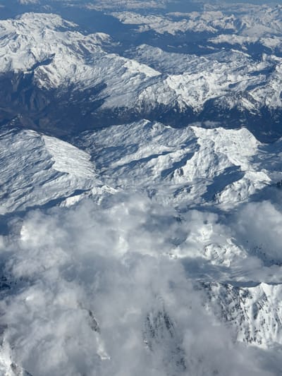 This aerial image depicts a vast, snow-covered mountain range in the French Alps, specifically within the vicinity of Le Monêtier-les-Bains. The scene is dominated by numerous rugged peaks and valleys, extensively blanketed in white snow, with darker, exposed rock or vegetated areas visible on some slopes and in lower elevations. A prominent valley with scattered, potentially built-up areas is discernible in the upper-left quadrant. Shadows cast by the elevated terrain suggest a high-angle light source, likely the sun, from the left. Layers of white and grey clouds partially obscure the lower and middle ground, drifting between peaks and valleys, with some mountains emerging above the cloud line. No identifiable people, explicit actions, or specific man-made objects beyond the implied settlement in the valley are present. The environment is a natural, high-altitude winter landscape.