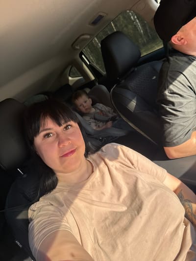 The image depicts the interior of a car, likely a selfie taken by a woman with dark hair and bangs. She is wearing a light-colored shirt. To the left of the woman is a child in a car seat, partially visible. Above them, the top of another person's head is visible, wearing a dark-colored cap and a dark-colored shirt. The car seems to have a sunroof. The location in Semenovo, Russia, cannot be confirmed from the image alone.