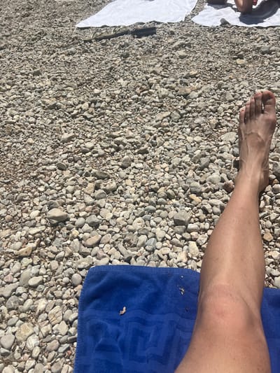 The image shows a gravel beach in Peguera, Spain. A person's lower leg and foot rest on the gravel, next to a blue towel. There is white fabric to the left of the frame.