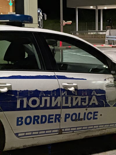 The image shows the side of a Serbian border police car. The Cyrillic letters "ГРАНИЧНА ПОЛИЦИЈА" are visible, along with "BORDER POLICE." The car is white with blue lettering. The scene appears to be at night, based on the lighting. The location is likely within Prijepolje Municipality, Serbia, as border police typically operate in border areas. Other details visible include a car window, door handles, and a side mirror. The car seems to be parked near some kind of control point structure with barriers and lights.