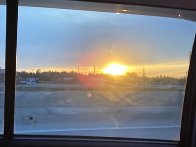 View from a car window with the setting sun visible over a highway in Bol'shiye Klyuchishchi, Russia. A forest and utility poles line the horizon behind the highway, and some parked vehicles are visible to the left. The highway barrier is in the foreground.