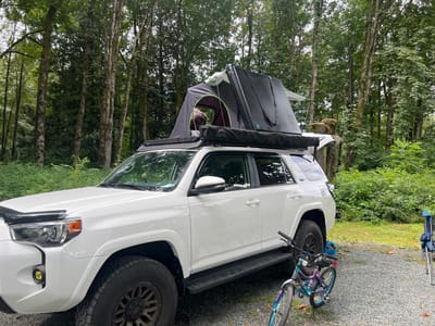 A white Toyota 4Runner is parked in a wooded area, likely a campsite, with a rooftop tent set up on its roof. A person wearing a plaid shirt is partially visible inside the tent. To the right of the vehicle, a bicycle with colorful accents is parked. In the background, numerous tall trees form a dense forest. A blue camping chair is visible in the far right of the frame. The ground is covered with gravel. The scene appears to be outdoors, in a natural setting. It is unclear if this location is specifically within the city of Chilliwack, Canada, as no identifying landmarks are present.