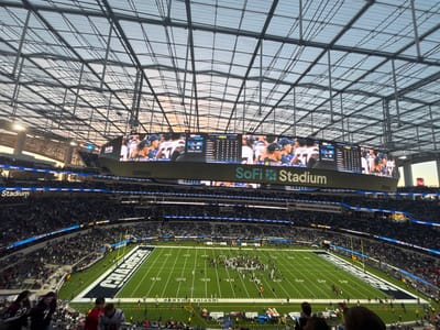 A football game at SoFi Stadium is underway. The Chargers' logo is clearly visible on the field. The stadium is packed with fans. The giant oval scoreboard displays images of the game. The stadium's distinctive roof structure is visible above. The sky appears to be early evening, with a hint of sunset.