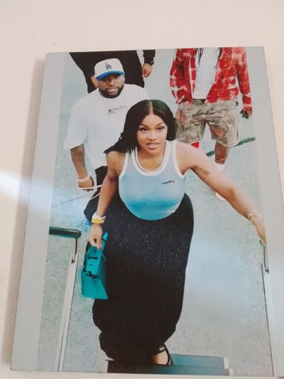 This media file features a photograph of three individuals ascending a set of stairs. In the foreground, a woman with dark hair and makeup is wearing a light blue tank top and a long, dark, sparkly skirt. She is carrying a turquoise handbag in her left hand and wears a yellow bracelet on her left wrist. She appears to be looking towards the camera. Behind her, a man in a white t-shirt and a blue and white baseball cap with "LA" written on it is walking up the stairs. He has a beard and is wearing a necklace. Further behind, another person is visible wearing a red and white patterned shirt and camouflage shorts. The setting appears to be indoors, possibly an airport or a similar public building, given the presence of stairs and the lighting. The overall mood of the photograph seems candid, capturing a moment of movement. No specific time of day or weather conditions are discernible from the image. There is text visible on the man's t-shirt, which appears to be a logo or design.