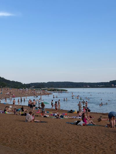This media file depicts a crowded beach on a sunny day, likely during the summer season. The scene is split between a vast expanse of clear blue sky and a beach filled with numerous people enjoying the water and sand. In the water, many individuals are swimming, wading, or floating, indicating a recreational day at the lake or sea. On the sandy shore, people are sunbathing, sitting on towels, or walking, suggesting a relaxed and leisurely atmosphere.
The background shows a tree-lined shore, hinting at a natural setting, possibly a lake within a forested area. The time of day appears to be afternoon or early evening, given the warm, soft lighting and the long shadows cast by some individuals on the sand. The weather seems pleasant, with clear skies and no visible indications of rain or wind. The overall mood is one of summer enjoyment and relaxation. There are no visible text elements in the image. The location context suggests this is a popular spot in the Ozyaritska-Slabadski rural council, Belarus.