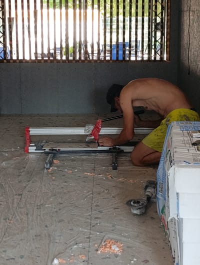 A shirtless person wearing a black cap and yellow shorts is kneeling on a construction site in Ladyzhyn, Ukraine, operating a tile cutter. The person is focused on the task, with their hands positioned on the cutter. The surrounding environment appears to be an unfinished building, with exposed walls and a floor covered in plastic sheeting. Construction materials, including what appear to be stacks of insulation, are visible in the foreground. Natural light filters in from a window or opening in the background, which is covered by vertical slats. An angle grinder is also present on the floor.