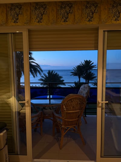 The image shows a view from inside a room, looking out onto a balcony and beyond to a sea or ocean landscape with palm trees at dusk or dawn. The location is likely Les Marines, Spain, given the Mediterranean vegetation and coastal scenery.
Inside, a window frame dominates the view, with an ornate, floral-patterned wall to the left and blinds visible in the upper and lower sections. The light indoors is dim, suggesting the outdoor light is the primary source.
On the balcony, there are rattan chairs and a glass-topped table. The balcony has a green railing and tiling is evident.
Beyond the balcony, the view extends to a body of water, likely the sea. The sky is a gradient of blue, indicative of the early morning or late evening. Palm trees are visible along the coastline, contributing to the Mediterranean aesthetic. The overall scene suggests a tranquil and relaxed atmosphere.