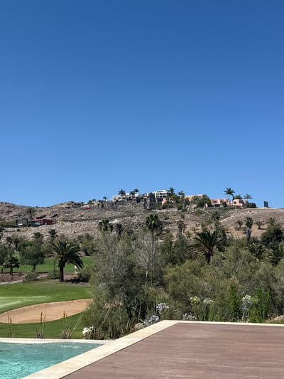 A wide shot captures a sunny day at Urbanización Salobre Golf, Spain. The foreground features a portion of a swimming pool with turquoise water and a wooden deck. Beyond this, lush green grass from a golf course is visible, including a sand trap. In the mid-ground, a dense arrangement of trees and shrubs, including palm trees, occupies the landscape. The background showcases a rocky hillside dotted with various buildings, some appearing residential, and more palm trees. The sky above is a clear, bright blue. No people are visible in the frame.