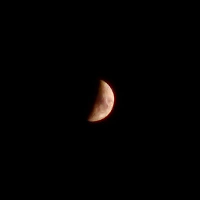 The image displays a waxing gibbous moon against a black, starless sky. The moon is positioned in the lower right quadrant of the frame. It is illuminated from the left, with the right side of the moon visible and catching the light. The surface of the moon shows discernible texture, with lighter and darker areas indicating craters and maria. The overall color of the illuminated portion is a warm, yellowish-orange hue, possibly due to atmospheric conditions or the camera's white balance. The image provides no information about the city of Calcinate, Italy, or any urban environment.