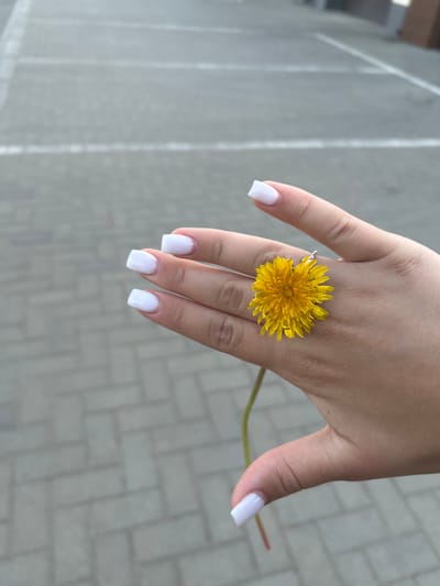 A close-up shot shows a human hand with white manicured nails holding a bright yellow dandelion flower. The flower is positioned so that the center of the blossom rests on the knuckle of the middle finger. A thin metal ring is visible on the ring finger. The background is a grey paved surface of rectangular blocks arranged in a herringbone pattern. Based on visual characteristics, it is not possible to confirm the location is the city of Chernomurovskii, Russia.