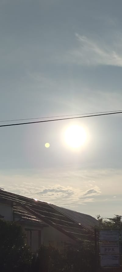 The image depicts a bright, sunny sky with some scattered clouds. The sun is a prominent feature, appearing as a large, bright orb in the upper right quadrant. To the left of the sun, a smaller, yellowish orb is visible, possibly a lens flare or another atmospheric phenomenon. A dark, horizontal power line stretches across the sky, crossing in front of the sun.
In the lower part of the image, the roof of a building is visible, angled from the left towards the center. The roof appears to be made of dark material and shows a pattern of horizontal beams or supports. The building itself is partially obscured by dark foliage in the foreground. To the right, a sign is visible, with text that is partially legible. The text includes "KOMITE" and "PPK", suggesting it is a sign related to a committee or organization.
The environment appears to be outdoors, likely in a semi-urban or rural setting given the presence of the building, power line, and foliage. The lighting suggests it is daytime, likely late morning or afternoon, due to the intensity of the sun.
The location is stated to be in Singgahmata, Indonesia. This context suggests the building and signage might be Indonesian in style and language.
