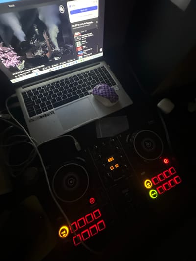 The image shows a dimly lit workspace with a laptop and DJ equipment. The laptop screen displays a YouTube video titled "JAY DE LYS @ Rio Electronico Music/La Plata 2024." A purple and white mouse rests on the laptop. To the right of the laptop, there is a DJ controller with illuminated buttons and dials, with a small white object nearby. Cables are visible connecting the laptop and the DJ equipment. In the bottom right corner is a CUE button, which is orange. The location is in La Plata, Argentina.