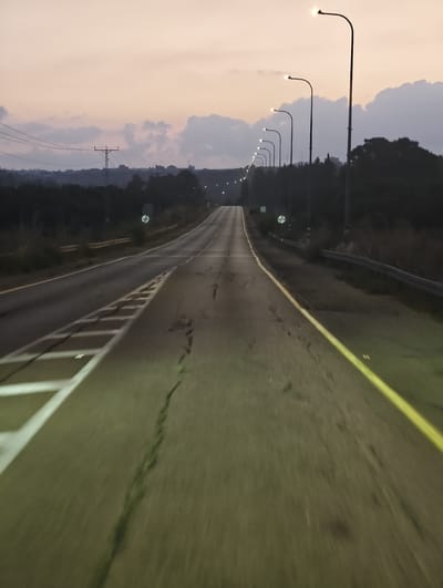 The image depicts a road in the vicinity of Horshim, Israel, during twilight or dawn. The road stretches into the distance, curving slightly to the left. The asphalt surface is worn and cracked in places, with a yellow line marking the right edge of the road and white lines delineating lanes and a merging area on the left. Streetlights, illuminated, line the right side of the road at regular intervals, casting a glow on the asphalt. The surrounding landscape is dark and appears to be covered in trees and brush. In the background, a few buildings are visible on a distant hill, and the sky is a pale orange and purple, with scattered clouds. The perspective suggests the photo was taken from a moving vehicle, indicated by the motion blur in the foreground. There are no people or animals visible in the scene. The overall atmosphere is one of solitude and a transition between day and night.