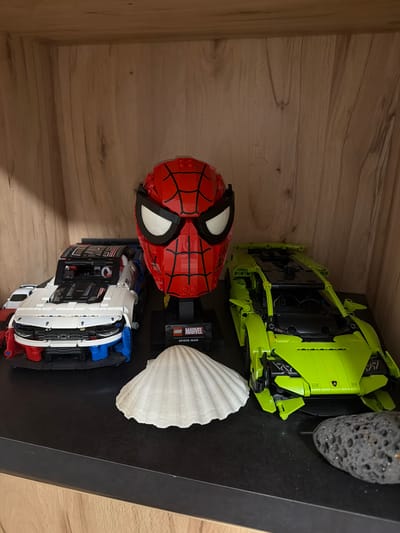 On a black shelf within a wooden cabinet, likely in Sankt Augustin, Germany, a collection of Lego models and natural objects are displayed. Prominently featured is a Lego Marvel Spider-Man helmet, a white Lego race car with red and blue accents, and a lime green Lego sports car. Between the Lego models sits a large, white scallop shell. To the right of the lime green car, a dark, porous rock rests on the shelf. The background consists of a light-colored wooden cabinet interior with visible grain.