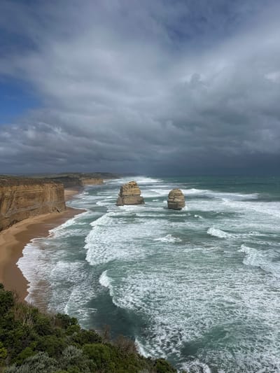 The image shows the Twelve Apostles rock formations in the ocean. The sky is filled with dark storm clouds contrasting with patches of blue. The ocean is rough, with large waves crashing against the shoreline and rock formations. A beach stretches along the coastline, and green vegetation grows on the edge of the cliffs. The scene conveys a sense of dramatic natural beauty, emphasizing the power of the ocean and the ruggedness of the coastal landscape.