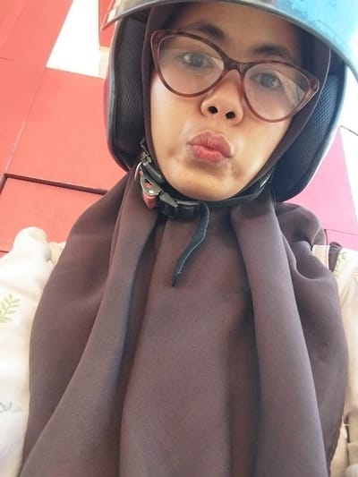 A medium-shot, close-up selfie of a young Indonesian woman wearing a blue helmet and a brown hijab, with her lips pursed in a kiss. She is wearing cat-eye glasses with brown frames. She is wearing a light-colored garment with a green leaf pattern peeking out from under her hijab. The background is a bright red wall. The scene appears to be outdoors in a public area, possibly near a building. The lighting suggests it is daytime.