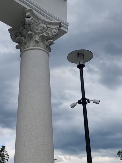 The image shows a close-up, low-angle view of a white classical column with ornate detailing at its capital, against a cloudy sky. The column is textured and appears to be made of stone or concrete. Attached to a dark pole extending horizontally across the frame are two white security cameras mounted on brackets. The pole also has a circular, flat, silver object attached to its left end, which could be a light fixture or a decorative element. The scene suggests an outdoor setting, possibly a public building or monument, given the column and security cameras. The time of day is uncertain, but the heavy clouds indicate overcast or approaching weather. There is no visible text in the image.