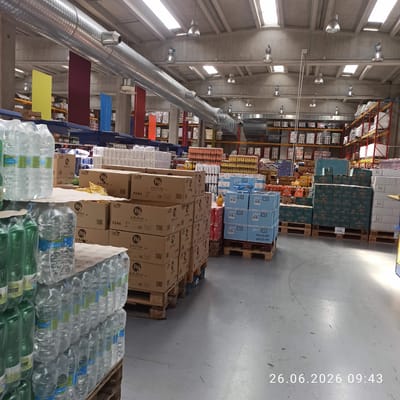 This image depicts the interior of a large warehouse or distribution center, likely a hypermarket or wholesale store, as suggested by the abundant shelves stocked with various products. The scene is dominated by rows of stacked boxes and pallets filled with goods. On the right side of the image, the floor is visible, marked with scuffs and faint lines, indicating a high-traffic area. The lighting suggests it is daytime, with natural light filtering in from high windows and artificial lights mounted on the ceiling and walls. The overall atmosphere is one of commercial activity and storage.
Visible text includes product labels and pricing information. On pallets in the foreground, we can see labels for "Pai patatine" and water bottles with brands like "GAVITA" and "FONTE NETTUNO". Further back, stacked boxes show text such as "GRANAROLO", "Colussi", and "OFFERTE". A prominent timestamp in the lower right corner reads "26.06.2026 09:43", indicating the date and time the photo was taken. The presence of "EPAL" markings on some pallets suggests they are European standard pallets, common in logistics.
The warehouse has a utilitarian aesthetic with exposed concrete walls and ceiling, along with large metal ventilation ducts running across the upper portion of the space. Various colored fabric or plastic sheets (red, blue, yellow) are suspended from the ceiling in the background, possibly for aesthetic purposes or to divide areas. There are no people visible in the image. The setting appears clean and organized, consistent with a functional retail or distribution environment.