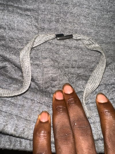 A grey drawstring with metal aglets lies on a grey fabric surface. A hand, dark-skinned, with short fingernails, rests partially on the fabric, near the drawstring. The fabric shows subtle texture. The drawstring is looped, creating a semi-circular shape. The aglets are a dark metal, contrasting with the grey of the drawstring. The lighting is uneven, creating shadows on the fabric and the hand.