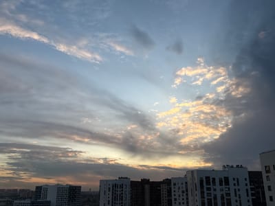 The image captures a cloudy sky over the city of Бордуки, Russia, likely during sunset or sunrise given the warm hues in the clouds. The sky is dominated by a dramatic display of clouds, with rays of sunlight breaking through in some areas, illuminating the edges of the clouds in shades of orange and yellow. To the right, the clouds are darker and more ominous, suggesting a weather front. In the foreground, several modern apartment buildings with numerous windows are visible, their architecture indicative of recent construction. The perspective suggests the photo was taken from a high vantage point, possibly another apartment building or a rooftop. The urban landscape extends into the distance, with more buildings visible on the horizon, blurring into a hazy silhouette against the sky. There are no people or discernible actions taking place in the image; it is a static scene focusing on the interplay of light and the urban environment.