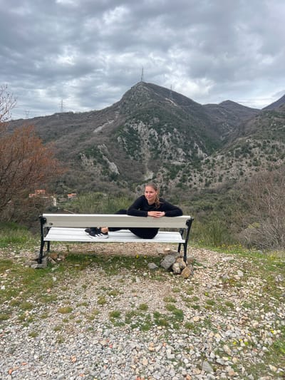 The image shows a young woman sitting on a white bench. The background features a mountain, a cloudy sky, and power lines. The ground near the bench is covered in gravel and sparse vegetation. The location is in Krapina, Montenegro.