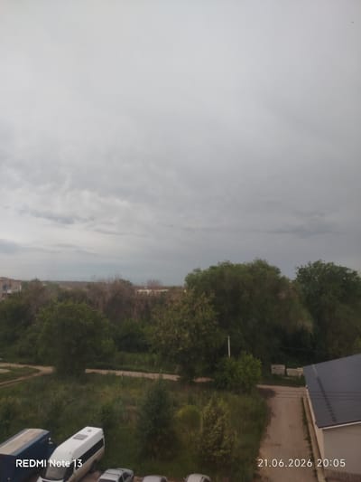 The image depicts an exterior view from a high vantage point, likely a window or balcony, looking towards a suburban or semi-rural landscape. The sky is overcast with gray clouds, suggesting it might be late afternoon or early evening, or that a storm is approaching. Below the sky, a line of trees and some buildings are visible.

In the foreground, on the left side of the image, there are several vehicles. A blue truck with a white container is parked, and next to it, a white van or bus is also parked. A silver car is partially visible in the lower right portion of the frame. The ground is covered with grass and vegetation, with a dirt road winding through the scene.

Further back, more trees and some low-rise buildings can be seen. The overall impression is one of a somewhat undeveloped or older residential area.

The bottom left corner of the image displays text that reads "REDMI Note 13". The bottom right corner shows the date and time: "21.06.2026 20:05". This indicates the photograph was taken on June 21, 2026, at 8:05 PM, and captured with a Redmi Note 13 device. The mention of Stroykeramika, Russia, provides a specific geographical context for the scene.