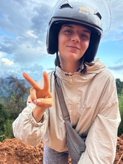 In this close-up selfie, a young woman wearing a dark grey helmet with a clear visor and a golden emblem that says "Royal" is pictured outdoors. She is looking directly at the camera with a faint smile, her right hand raised in a peace sign. She is wearing a light beige hooded jacket with a zipper and a grey strap bag slung across her body. The background shows a cloudy sky with patches of blue and hints of greenery from trees and foliage, suggesting a natural or rural setting, possibly in or around Nao Sẻ, Vietnam. The terrain in the foreground appears to be made of reddish-brown soil. The helmet has visible water droplets on it, indicating recent rain or high humidity.