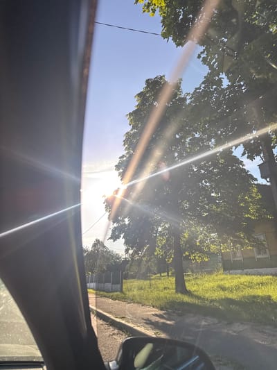 The image captures a sun-drenched scene, likely from a moving vehicle in the vicinity of Азгінавіцкі сельскі Савет, Belarus. The sun, a brilliant source of light, creates prominent lens flares and streaks across the frame, obscuring some details. In the foreground, the dark interior of a vehicle is visible, with a portion of the windshield and possibly a side pillar. Beyond the vehicle, a paved path or road runs alongside a grassy verge. Further into the background, lush green trees dominate the landscape, their leaves illuminated by the sunlight. A traditional wooden house with a yellow facade and white trim is partially visible behind the trees. The sky is a clear, bright blue, with a few wispy clouds. The overall impression is one of a bright, summer day in a rural or semi-rural setting.