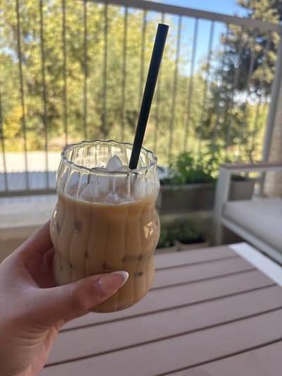 The image depicts a close-up, outdoor scene located in Maale Adumim, Palestinian Territory, likely on a balcony or patio. The primary subject is a clear, rippled glass holding an iced coffee beverage with a black straw. The glass contains a light brown, milky liquid with numerous ice cubes visible, some partially melted, and condensation on its exterior. A left hand, with manicured nails featuring a French tip design (pink base with white tips), grips the lower portion of the glass. The hand and glass are positioned in the foreground, slightly right of center.
In the mid-ground, to the right, a light-colored slatted table or surface is partially visible. Further to the right, a section of modern outdoor furniture with light-colored cushions is present. The background features a blurred natural environment with abundant green foliage, indicative of trees or shrubs, under daylight conditions. A dark metal railing runs horizontally across the upper mid-ground, separating the immediate area from the distant greenery. The sky, visible in the upper right, is bright blue.